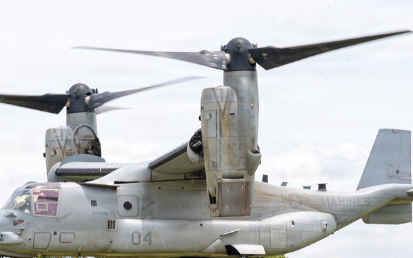 A U.S. Marines MV-22 Osprey tiltrotor aircraft (bureau number 8033, side number 04) hovers at low altitude over a grassy field. A soldier in camouflage uniform stands beneath or near the aircraft next to what appears to be an artillery piece or heavy weapon system. The scene suggests a military training exercise or sling-load/fire support coordination operation in a tropical or subtropical environment.