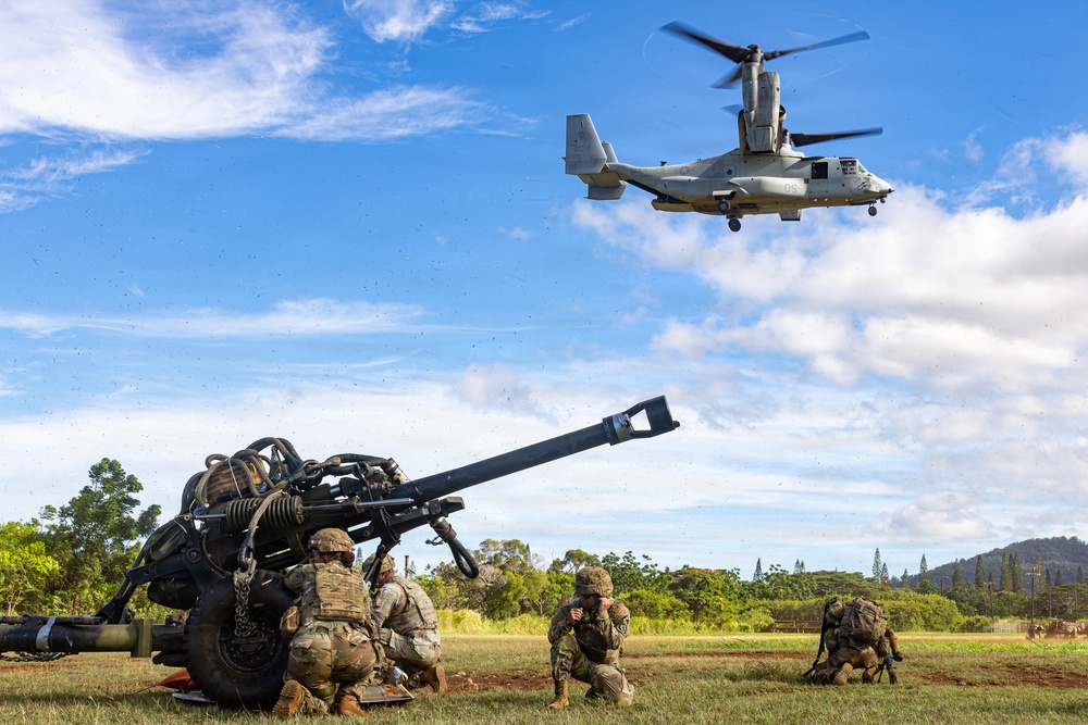 Military personnel in camouflage uniforms are positioned around what appears to be an M777 lightweight howitzer artillery piece on an open grassy field. A V-22 Osprey tiltrotor aircraft, marked with the number '05', is hovering low overhead, likely conducting a sling-load or air assault exercise. The setting appears to be a tropical or subtropical training area, consistent with locations such as Hawaii.