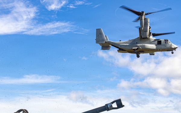 Military personnel in camouflage uniforms are positioned around what appears to be an M777 lightweight howitzer artillery piece on an open grassy field. A V-22 Osprey tiltrotor aircraft, marked with the number '05', is hovering low overhead, likely conducting a sling-load or air assault exercise. The setting appears to be a tropical or subtropical training area, consistent with locations such as Hawaii.