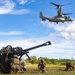 Military personnel in camouflage uniforms are positioned around what appears to be an M777 lightweight howitzer artillery piece on an open grassy field. A V-22 Osprey tiltrotor aircraft, marked with the number '05', is hovering low overhead, likely conducting a sling-load or air assault exercise. The setting appears to be a tropical or subtropical training area, consistent with locations such as Hawaii.