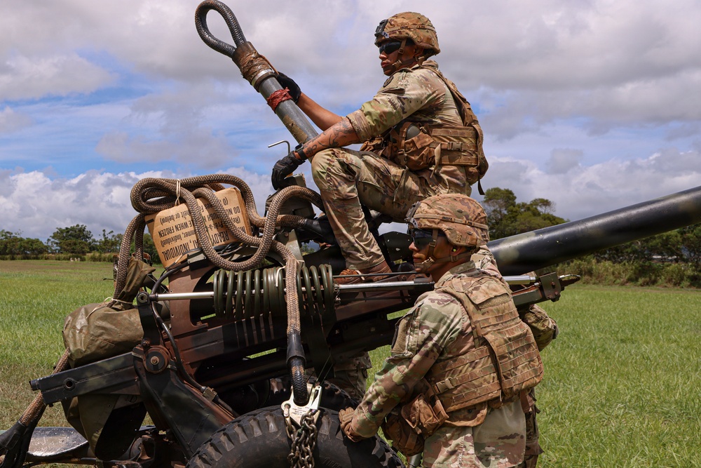 Two soldiers in U.S. Army Operational Camouflage Pattern (OCP) uniforms are working on what appears to be an M777 lightweight howitzer or similar artillery piece in an open grassy field. One soldier is positioned on top of the equipment while the other assists from ground level. The setting appears to be a training exercise based on the lush tropical vegetation visible in the background, consistent with a location such as Hawaii.