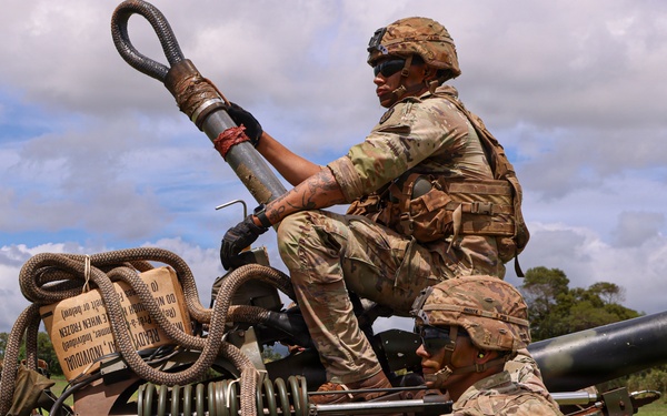 Two soldiers in U.S. Army Operational Camouflage Pattern (OCP) uniforms are working on what appears to be an M777 lightweight howitzer or similar artillery piece in an open grassy field. One soldier is positioned on top of the equipment while the other assists from ground level. The setting appears to be a training exercise based on the lush tropical vegetation visible in the background, consistent with a location such as Hawaii.