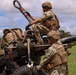 Two soldiers in U.S. Army Operational Camouflage Pattern (OCP) uniforms are working on what appears to be an M777 lightweight howitzer or similar artillery piece in an open grassy field. One soldier is positioned on top of the equipment while the other assists from ground level. The setting appears to be a training exercise based on the lush tropical vegetation visible in the background, consistent with a location such as Hawaii.