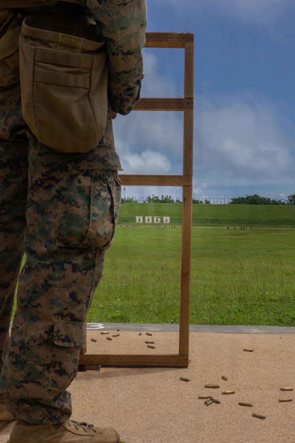 A military personnel in MARPAT digital camouflage uniform stands at a firing line on an outdoor shooting range. Multiple spent brass casings are visible on the ground, indicating recent firing activity. Silhouette targets are positioned downrange on a grassy berm under a partly cloudy sky.