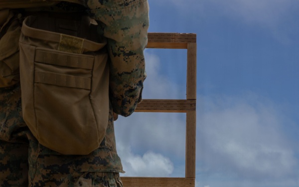 A military personnel in MARPAT digital camouflage uniform stands at a firing line on an outdoor shooting range. Multiple spent brass casings are visible on the ground, indicating recent firing activity. Silhouette targets are positioned downrange on a grassy berm under a partly cloudy sky.