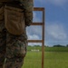 A military personnel in MARPAT digital camouflage uniform stands at a firing line on an outdoor shooting range. Multiple spent brass casings are visible on the ground, indicating recent firing activity. Silhouette targets are positioned downrange on a grassy berm under a partly cloudy sky.