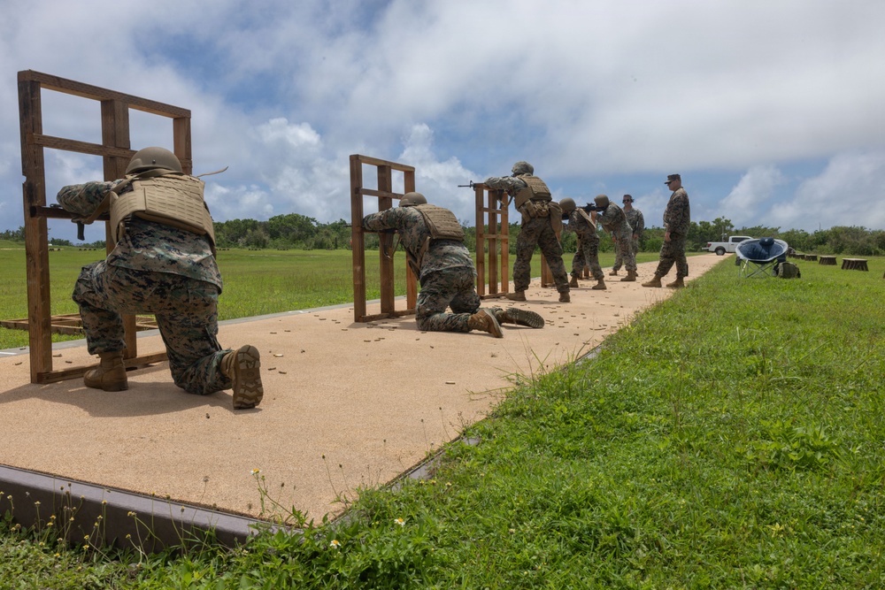 A group of military personnel in MARPAT camouflage uniforms are conducting a live-fire rifle range exercise on a shooting platform. Multiple shooters are positioned at wooden barricades in various firing stances, including kneeling and standing, while an instructor or range safety officer observes from the right. Spent brass casings are visible on the sandy platform surface, indicating active firing.