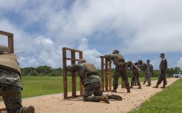 A group of military personnel in MARPAT camouflage uniforms are conducting a live-fire rifle range exercise on a shooting platform. Multiple shooters are positioned at wooden barricades in various firing stances, including kneeling and standing, while an instructor or range safety officer observes from the right. Spent brass casings are visible on the sandy platform surface, indicating active firing.
