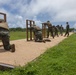 A group of military personnel in MARPAT camouflage uniforms are conducting a live-fire rifle range exercise on a shooting platform. Multiple shooters are positioned at wooden barricades in various firing stances, including kneeling and standing, while an instructor or range safety officer observes from the right. Spent brass casings are visible on the sandy platform surface, indicating active firing.