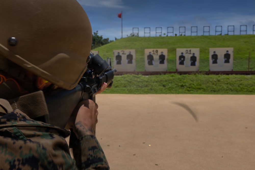 A military service member in camouflage uniform and tactical helmet is aiming a rifle at a shooting range. Multiple human silhouette targets are visible downrange, numbered and mounted on stands in front of a grass berm. A red range flag is visible on a pole in the background, indicating an active firing range.