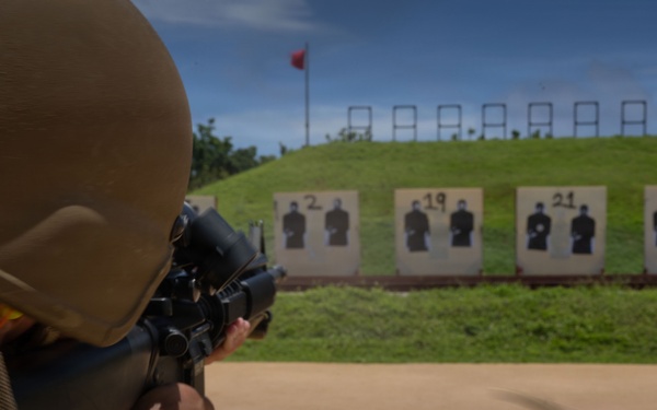 A military service member in camouflage uniform and tactical helmet is aiming a rifle at a shooting range. Multiple human silhouette targets are visible downrange, numbered and mounted on stands in front of a grass berm. A red range flag is visible on a pole in the background, indicating an active firing range.