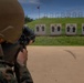 A military service member in camouflage uniform and tactical helmet is aiming a rifle at a shooting range. Multiple human silhouette targets are visible downrange, numbered and mounted on stands in front of a grass berm. A red range flag is visible on a pole in the background, indicating an active firing range.