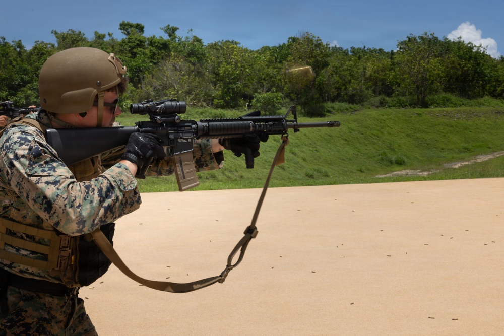 A military service member in MARPAT digital camouflage uniform is actively firing an M16/M4-style rifle at an outdoor shooting range. A spent brass casing is visibly ejecting from the weapon, indicating the moment of discharge. Additional personnel in similar uniforms are visible in the background, suggesting a group training exercise.