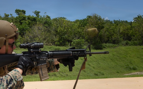 A military service member in MARPAT digital camouflage uniform is actively firing an M16/M4-style rifle at an outdoor shooting range. A spent brass casing is visibly ejecting from the weapon, indicating the moment of discharge. Additional personnel in similar uniforms are visible in the background, suggesting a group training exercise.