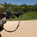 A military service member in MARPAT digital camouflage uniform is actively firing an M16/M4-style rifle at an outdoor shooting range. A spent brass casing is visibly ejecting from the weapon, indicating the moment of discharge. Additional personnel in similar uniforms are visible in the background, suggesting a group training exercise.