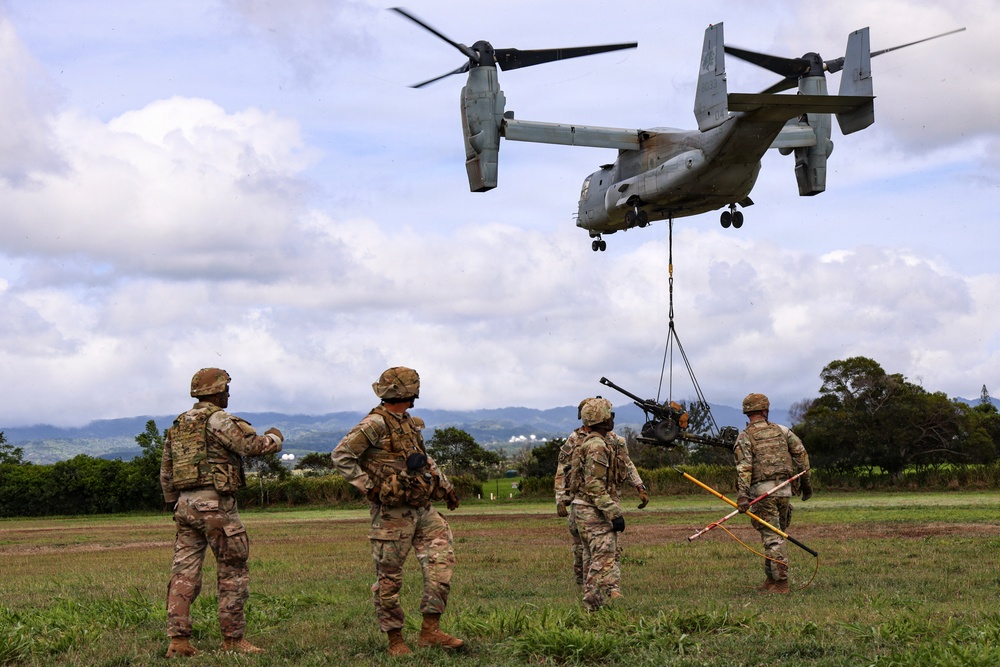 A military tiltrotor aircraft, identified as a Bell Boeing V-22 Osprey, hovers low over a grassy field while conducting a sling-load operation, lifting what appears to be an artillery piece or military equipment. Four soldiers in MultiCam/OCP camouflage uniforms and full combat gear observe and guide the operation from the ground. The setting appears to be a tropical or subtropical training area with green hills and scattered trees in the background.