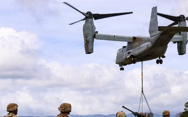 A military tiltrotor aircraft, identified as a Bell Boeing V-22 Osprey, hovers low over a grassy field while conducting a sling-load operation, lifting what appears to be an artillery piece or military equipment. Four soldiers in MultiCam/OCP camouflage uniforms and full combat gear observe and guide the operation from the ground. The setting appears to be a tropical or subtropical training area with green hills and scattered trees in the background.