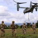 A military tiltrotor aircraft, identified as a Bell Boeing V-22 Osprey, hovers low over a grassy field while conducting a sling-load operation, lifting what appears to be an artillery piece or military equipment. Four soldiers in MultiCam/OCP camouflage uniforms and full combat gear observe and guide the operation from the ground. The setting appears to be a tropical or subtropical training area with green hills and scattered trees in the background.