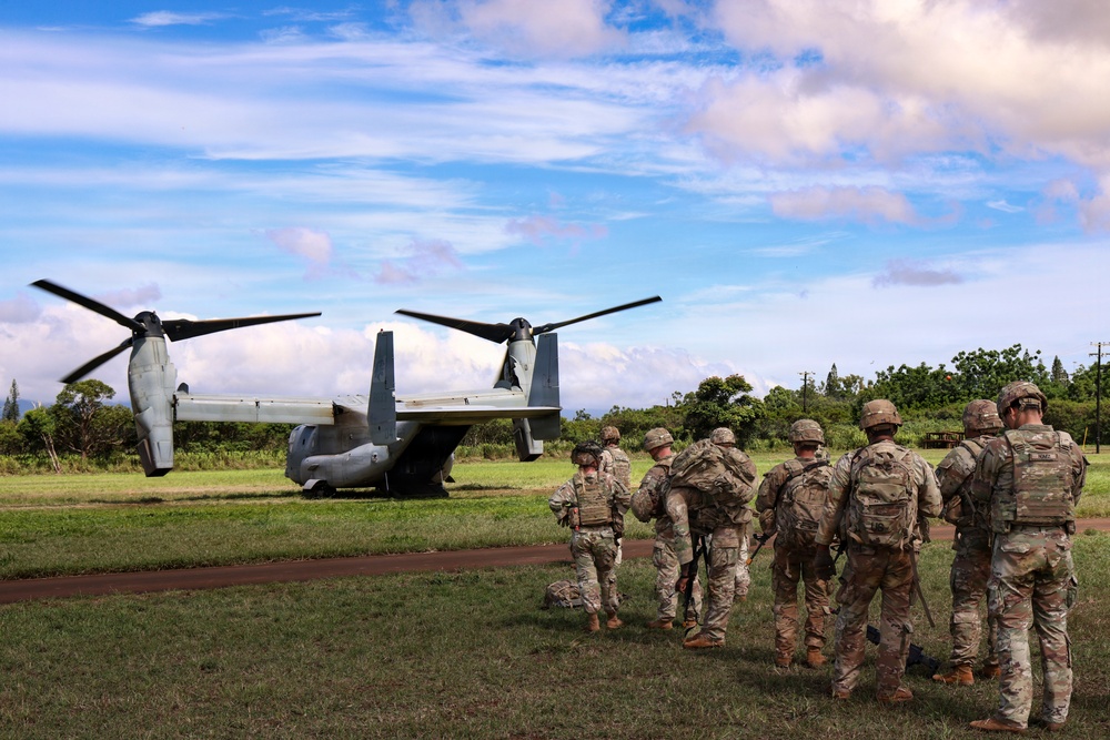 A group of military personnel in camouflage uniforms are gathered near a V-22 Osprey tiltrotor aircraft on a grassy field. The soldiers appear to be preparing for or debriefing after an air assault or insertion operation. The setting appears to be a tropical or subtropical environment based on the lush vegetation and cloud formations.