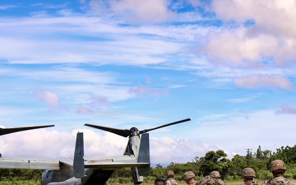 A group of military personnel in camouflage uniforms are gathered near a V-22 Osprey tiltrotor aircraft on a grassy field. The soldiers appear to be preparing for or debriefing after an air assault or insertion operation. The setting appears to be a tropical or subtropical environment based on the lush vegetation and cloud formations.