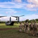A group of military personnel in camouflage uniforms are gathered near a V-22 Osprey tiltrotor aircraft on a grassy field. The soldiers appear to be preparing for or debriefing after an air assault or insertion operation. The setting appears to be a tropical or subtropical environment based on the lush vegetation and cloud formations.
