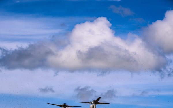 A military tiltrotor aircraft, identifiable as a Bell Boeing V-22 Osprey, is hovering or landing over a grassy field with red-dirt paths. The aircraft's rotors are tilted upward in helicopter mode, kicking up significant debris and dust from the ground below. The setting appears to be a rural or training area with dense tree lines and hills in the background under a partly cloudy sky.