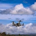 A military tiltrotor aircraft, identifiable as a Bell Boeing V-22 Osprey, is hovering or landing over a grassy field with red-dirt paths. The aircraft's rotors are tilted upward in helicopter mode, kicking up significant debris and dust from the ground below. The setting appears to be a rural or training area with dense tree lines and hills in the background under a partly cloudy sky.