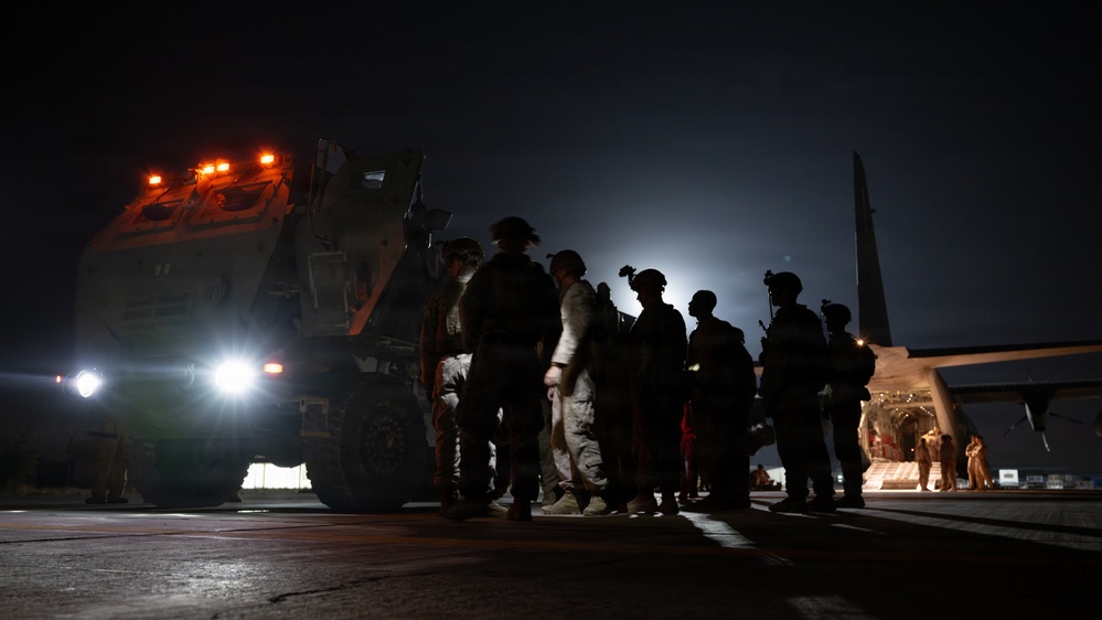 A nighttime military operation on what appears to be an airfield or tarmac. Several soldiers in combat gear are gathered near a large military wheeled vehicle with orange warning lights illuminated, adjacent to what appears to be a military transport aircraft with its rear ramp open. The scene is predominantly dark with artificial lighting from vehicle headlights and warning lights.