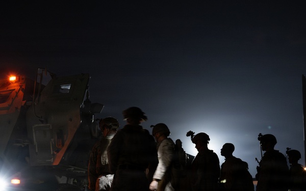 A nighttime military operation on what appears to be an airfield or tarmac. Several soldiers in combat gear are gathered near a large military wheeled vehicle with orange warning lights illuminated, adjacent to what appears to be a military transport aircraft with its rear ramp open. The scene is predominantly dark with artificial lighting from vehicle headlights and warning lights.