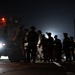 A nighttime military operation on what appears to be an airfield or tarmac. Several soldiers in combat gear are gathered near a large military wheeled vehicle with orange warning lights illuminated, adjacent to what appears to be a military transport aircraft with its rear ramp open. The scene is predominantly dark with artificial lighting from vehicle headlights and warning lights.