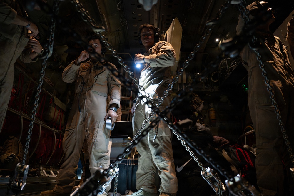 Two individuals wearing tan/khaki flight suits and aviation headsets are standing inside what appears to be a military cargo aircraft. The interior is dimly lit, with heavy chains in the foreground used for securing cargo. One person is holding a flashlight while the other appears to be communicating via headset.