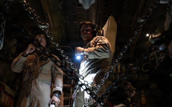 Two individuals wearing tan/khaki flight suits and aviation headsets are standing inside what appears to be a military cargo aircraft. The interior is dimly lit, with heavy chains in the foreground used for securing cargo. One person is holding a flashlight while the other appears to be communicating via headset.