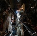 Two individuals wearing tan/khaki flight suits and aviation headsets are standing inside what appears to be a military cargo aircraft. The interior is dimly lit, with heavy chains in the foreground used for securing cargo. One person is holding a flashlight while the other appears to be communicating via headset.