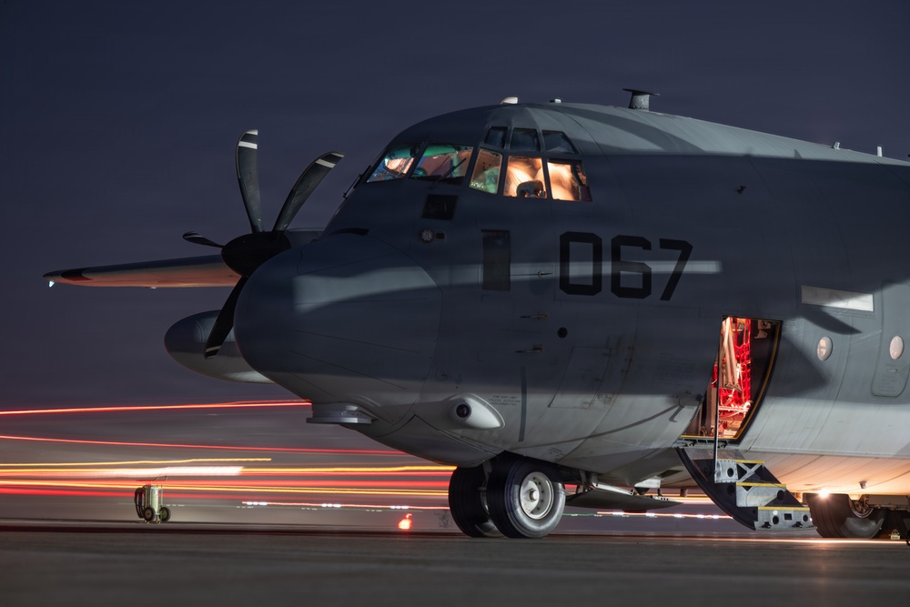 A military turboprop aircraft marked with the number '067' is photographed on a tarmac at night. The cockpit is illuminated from within, and a side door is open with red interior lighting visible. Light trails from ground vehicles streak across the tarmac in the background, suggesting long-exposure photography.
