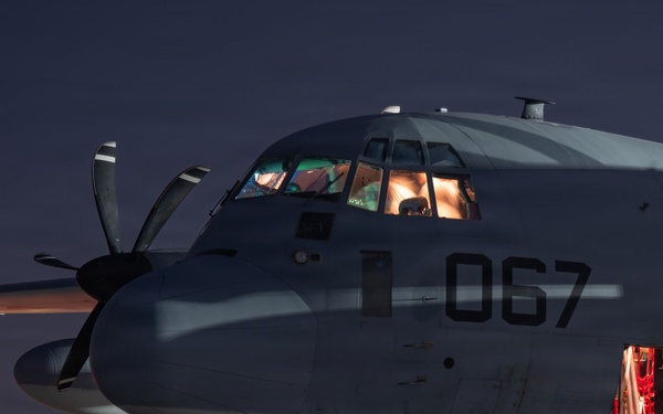 A military turboprop aircraft marked with the number '067' is photographed on a tarmac at night. The cockpit is illuminated from within, and a side door is open with red interior lighting visible. Light trails from ground vehicles streak across the tarmac in the background, suggesting long-exposure photography.
