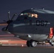 A military turboprop aircraft marked with the number '067' is photographed on a tarmac at night. The cockpit is illuminated from within, and a side door is open with red interior lighting visible. Light trails from ground vehicles streak across the tarmac in the background, suggesting long-exposure photography.
