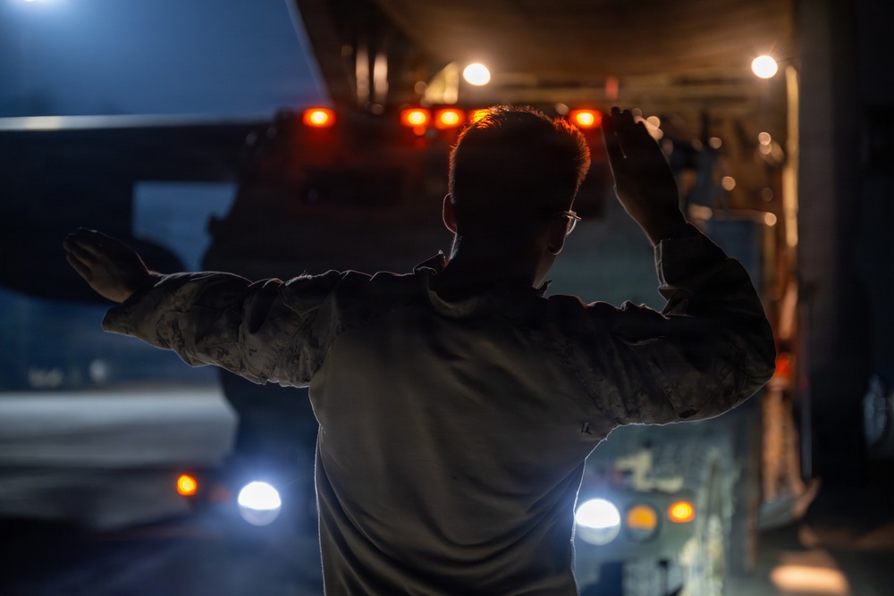 A person in military camouflage uniform is directing a large vehicle at night, using hand signals with both arms raised. The scene is illuminated by orange and white vehicle lights, creating a dramatic low-light environment. The individual appears to be a ground guide or marshal directing the movement of a large military or heavy transport vehicle.