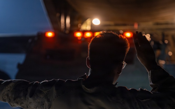 A person in military camouflage uniform is directing a large vehicle at night, using hand signals with both arms raised. The scene is illuminated by orange and white vehicle lights, creating a dramatic low-light environment. The individual appears to be a ground guide or marshal directing the movement of a large military or heavy transport vehicle.