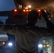 A person in military camouflage uniform is directing a large vehicle at night, using hand signals with both arms raised. The scene is illuminated by orange and white vehicle lights, creating a dramatic low-light environment. The individual appears to be a ground guide or marshal directing the movement of a large military or heavy transport vehicle.