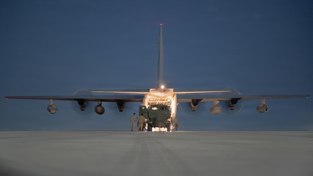 A large military transport aircraft, consistent with a C-130 Hercules, is photographed from the rear at dusk or dawn on a tarmac or airstrip. The aircraft's rear cargo ramp is open and illuminated, with a military vehicle being loaded or unloaded. Several personnel are visible near the rear of the aircraft overseeing the operation.