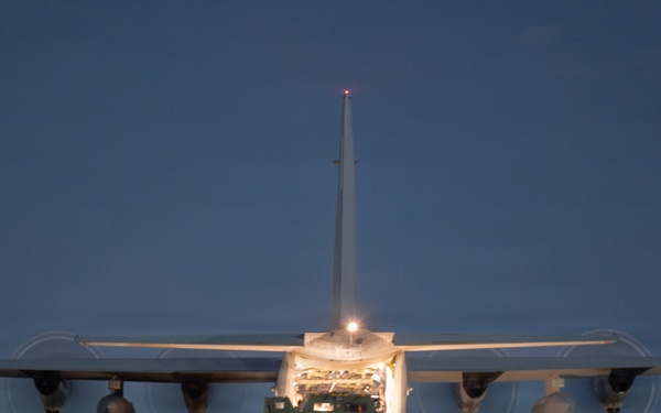 A large military transport aircraft, consistent with a C-130 Hercules, is photographed from the rear at dusk or dawn on a tarmac or airstrip. The aircraft's rear cargo ramp is open and illuminated, with a military vehicle being loaded or unloaded. Several personnel are visible near the rear of the aircraft overseeing the operation.