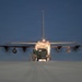 A large military transport aircraft, consistent with a C-130 Hercules, is photographed from the rear at dusk or dawn on a tarmac or airstrip. The aircraft's rear cargo ramp is open and illuminated, with a military vehicle being loaded or unloaded. Several personnel are visible near the rear of the aircraft overseeing the operation.