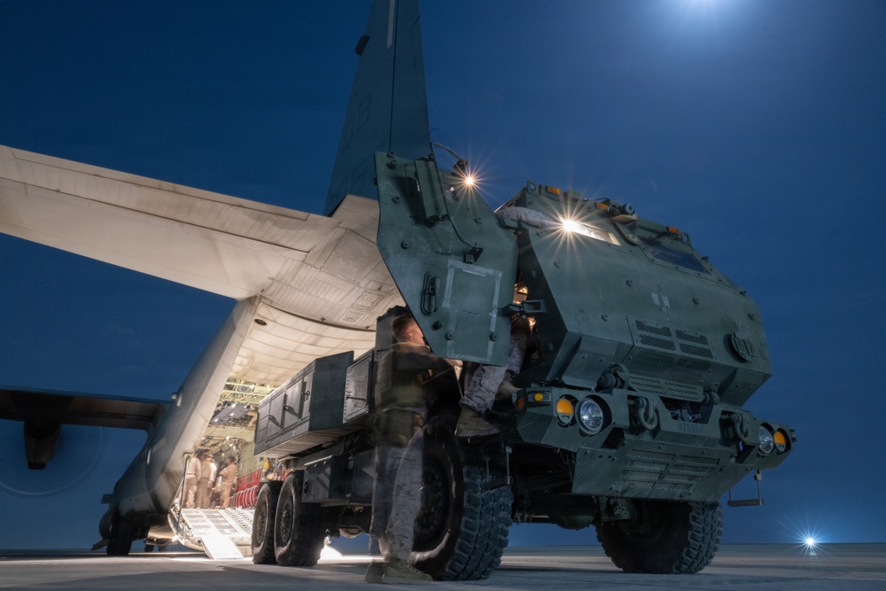 A military ground vehicle, appearing to be an HIMARS (High Mobility Artillery Rocket System) or similar wheeled rocket launcher, is being loaded onto a large military transport aircraft at night. The operation is taking place on an airfield tarmac under a deep blue night sky with a visible moon. Personnel in military uniforms are guiding the vehicle up the aircraft's rear loading ramp.
