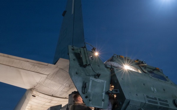 A military ground vehicle, appearing to be an HIMARS (High Mobility Artillery Rocket System) or similar wheeled rocket launcher, is being loaded onto a large military transport aircraft at night. The operation is taking place on an airfield tarmac under a deep blue night sky with a visible moon. Personnel in military uniforms are guiding the vehicle up the aircraft's rear loading ramp.