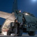 A military ground vehicle, appearing to be an HIMARS (High Mobility Artillery Rocket System) or similar wheeled rocket launcher, is being loaded onto a large military transport aircraft at night. The operation is taking place on an airfield tarmac under a deep blue night sky with a visible moon. Personnel in military uniforms are guiding the vehicle up the aircraft's rear loading ramp.