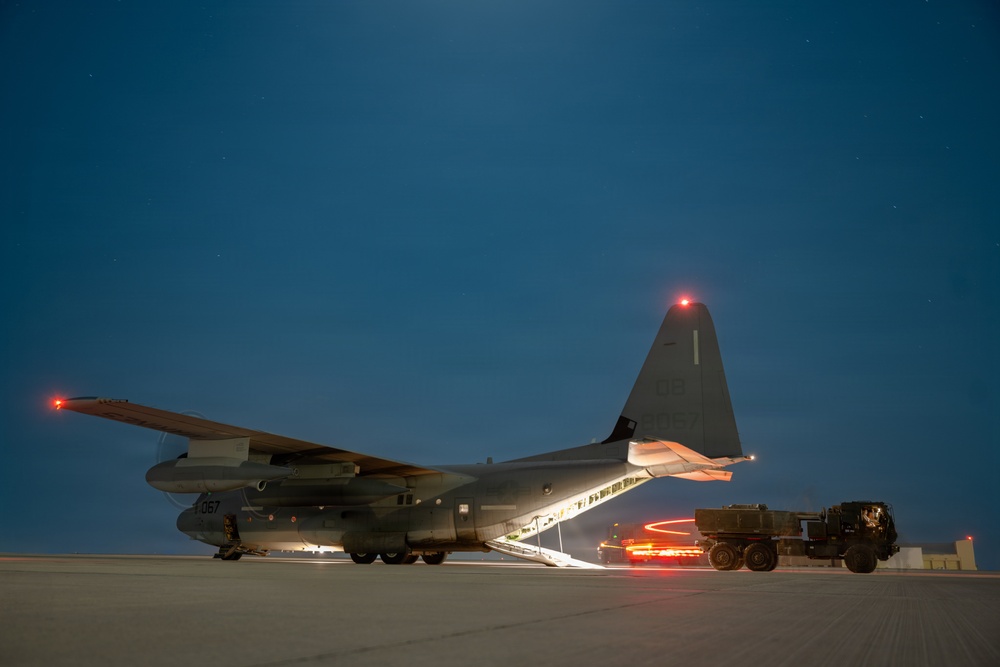 A military transport aircraft, identifiable as a C-130 Hercules with tail number ending in '067', is photographed on a tarmac at night or dusk. The rear cargo ramp is lowered and illuminated, with a military truck positioned at the ramp, suggesting active loading or unloading operations. Navigation lights are visible on the wingtip and tail, and the deep blue twilight sky provides a dramatic backdrop.