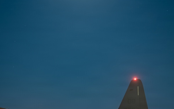 A military transport aircraft, identifiable as a C-130 Hercules with tail number ending in '067', is photographed on a tarmac at night or dusk. The rear cargo ramp is lowered and illuminated, with a military truck positioned at the ramp, suggesting active loading or unloading operations. Navigation lights are visible on the wingtip and tail, and the deep blue twilight sky provides a dramatic backdrop.
