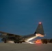 A military transport aircraft, identifiable as a C-130 Hercules with tail number ending in '067', is photographed on a tarmac at night or dusk. The rear cargo ramp is lowered and illuminated, with a military truck positioned at the ramp, suggesting active loading or unloading operations. Navigation lights are visible on the wingtip and tail, and the deep blue twilight sky provides a dramatic backdrop.