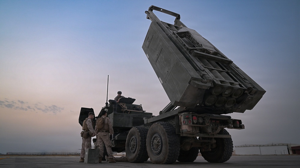 The image shows a multiple launch rocket system (MLRS) mounted on a heavy military truck with its launcher pod elevated at an angle. Several military personnel in camouflage uniforms are gathered around the vehicle, apparently conducting operations or maintenance. The scene appears to take place on a paved surface during dusk or dawn conditions.