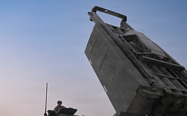 The image shows a multiple launch rocket system (MLRS) mounted on a heavy military truck with its launcher pod elevated at an angle. Several military personnel in camouflage uniforms are gathered around the vehicle, apparently conducting operations or maintenance. The scene appears to take place on a paved surface during dusk or dawn conditions.